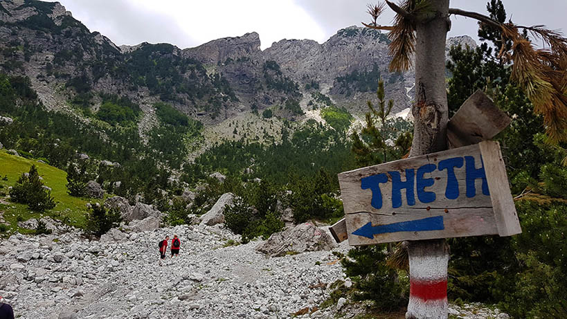 Wooden sign on a path in the Albanian Alps pointing left with Thethi painted on it with blue paint.